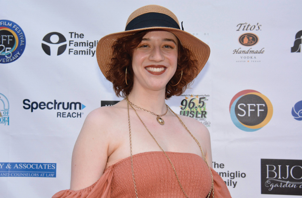 Acacia DëQueer, class of 2019, poses in a hat and summery dress in front of a step-and-repeat at the premiere of a film.