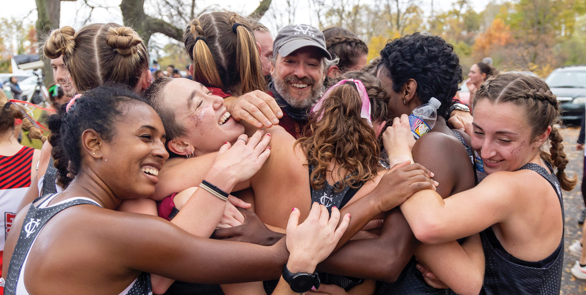 A group of young women cross country runners, hug each other in celebration as they crowd around their coach at center.