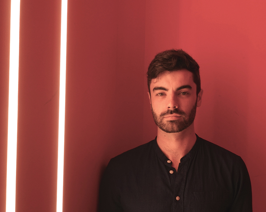 A portrait of Will Healy, class of 2012, with a red background, standing next to two cylindrical lights.