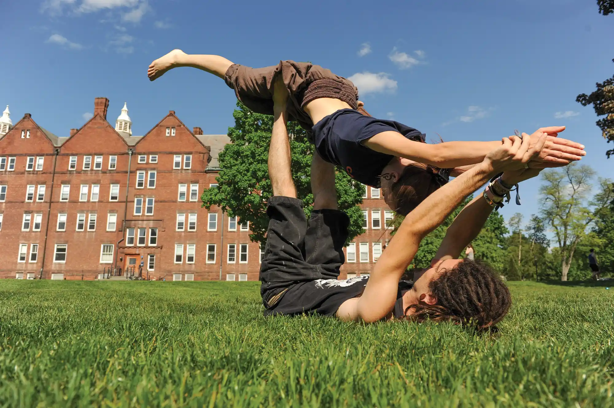 A male student on his back on the grassy Quad playfully lifts a female student in the air with his feet.