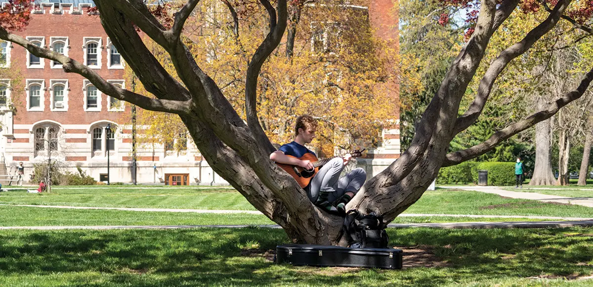 A male student plays a guitar as he sits at the base of a tree on the Quad.