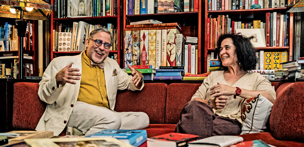 Professor Epstein and his spouse, Professor Rabbanit Agit Veto, sit on a red couch in front of a bookshelf, laughing together.