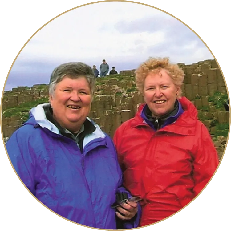 Two women smiling outdoors, one wearing a purple jacket and the other a red jacket, standing in front of a rock formation under a cloudy sky.