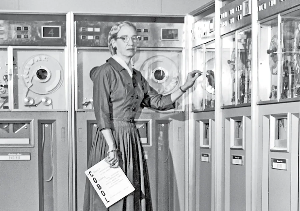 A black-and-white photo of a woman in a dress and glasses standing in front of a wall of early computer equipment. She holds a paper labeled "COBOL" while adjusting one of the computer controls.