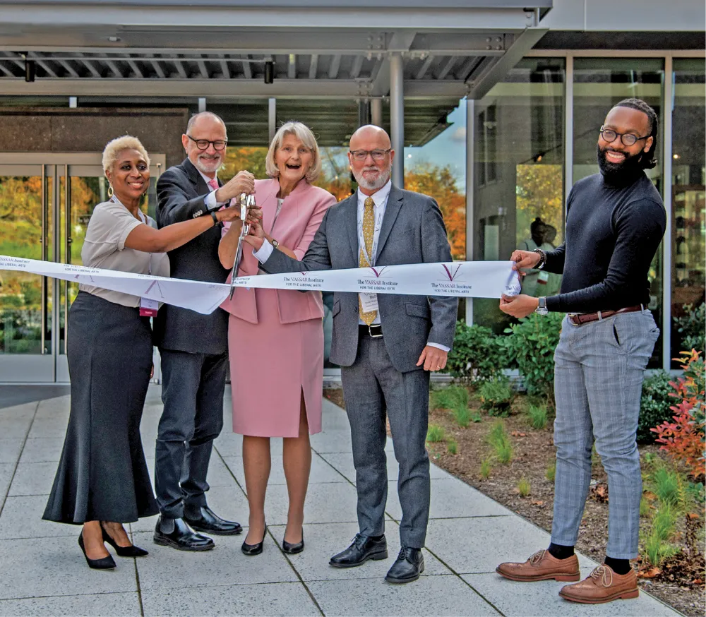 Five people stand in front of a modern glass building holding a ribbon-cutting ceremony. The group includes two women and three men, one of whom holds scissors. They are all dressed in formal attire and smiling.