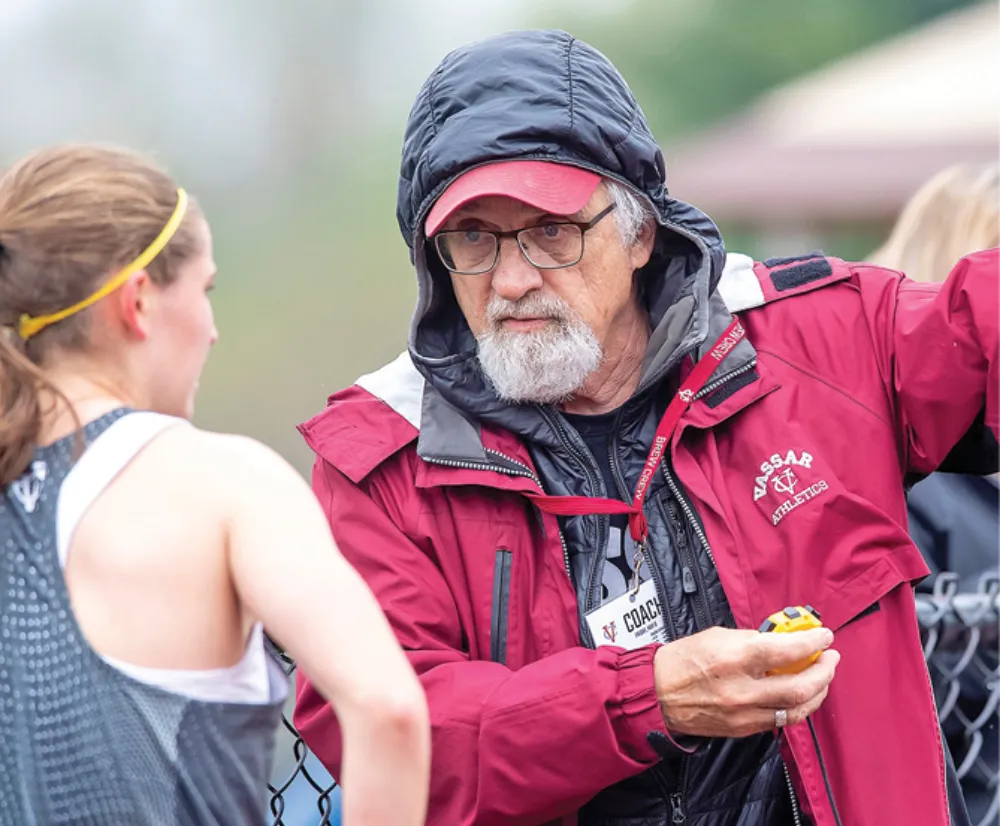 A coach in a maroon Vassar Athletics jacket and black hoodie speaks to a female athlete in a gray uniform with a yellow headband. The coach is holding a stopwatch and leans on a fence.