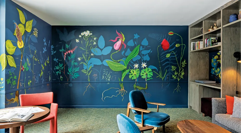 A colorful mural of oversized botanical plants decorates a deep blue wall in a cozy sitting area. The room includes bookshelves, modern chairs, a wooden table, and a red chair in the foreground.