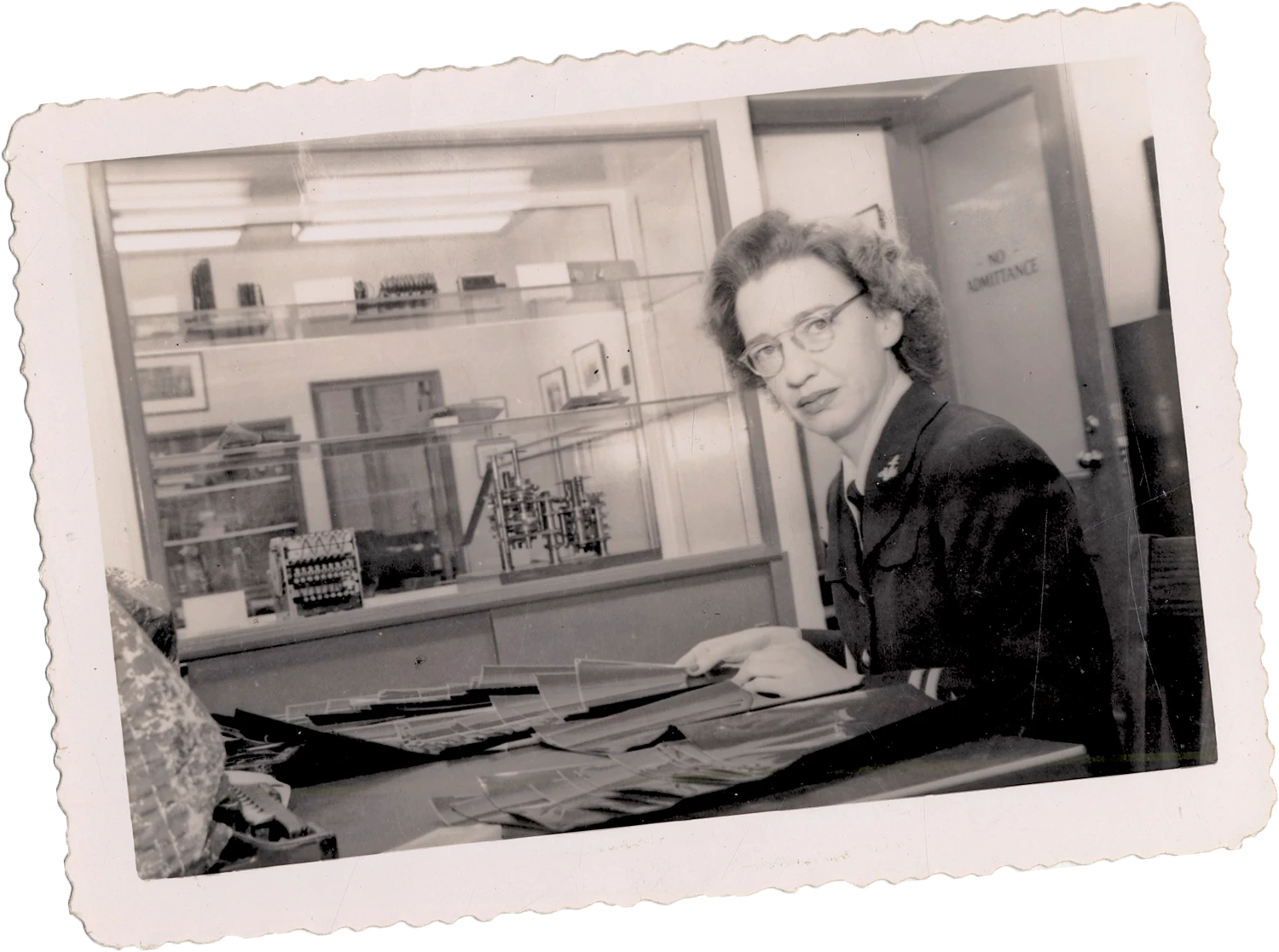 A black-and-white photo of a woman in glasses, seated at a desk in an office or lab with technical equipment in the background.