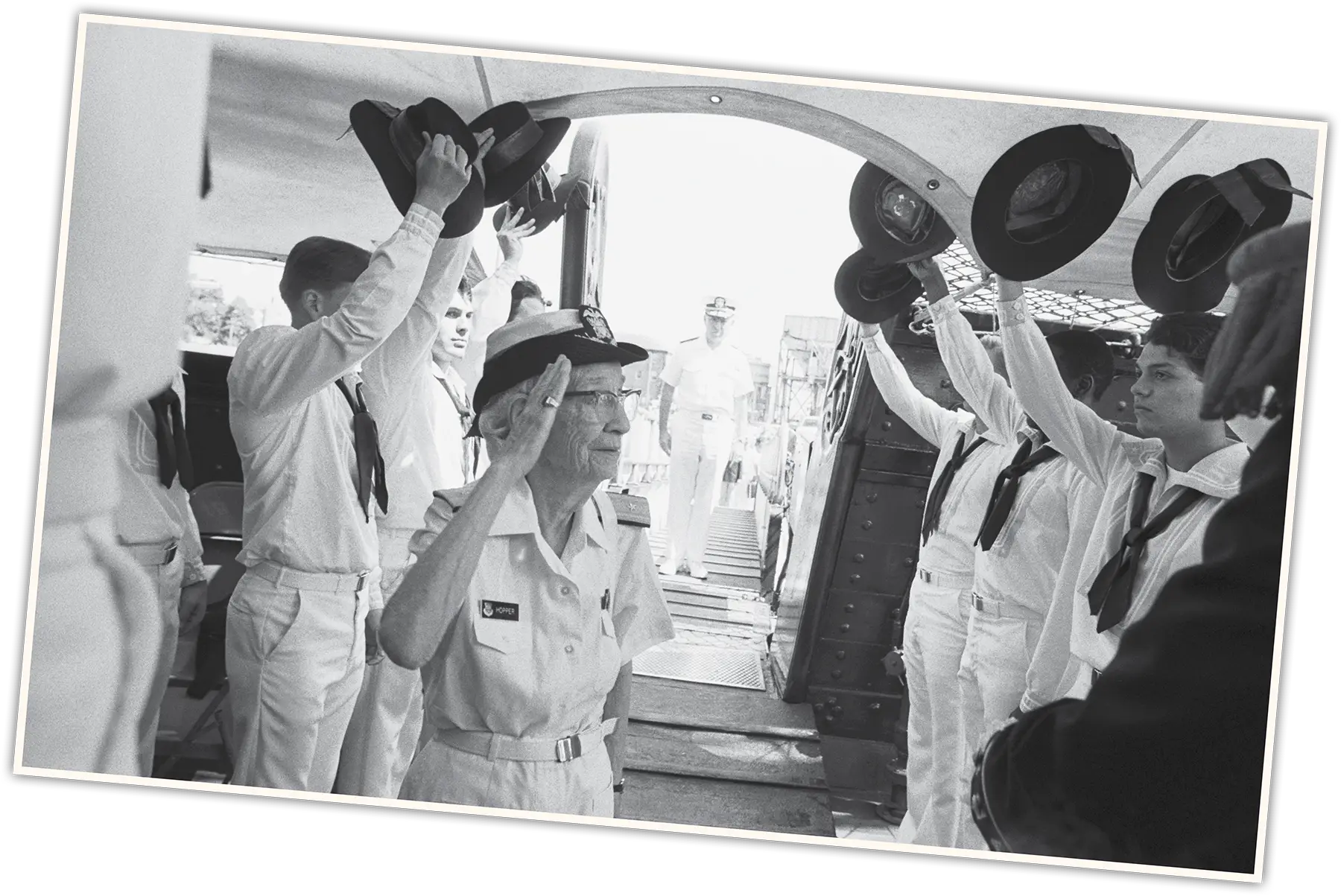 A woman in naval uniform salutes while walking under an archway formed by officers holding up hats.