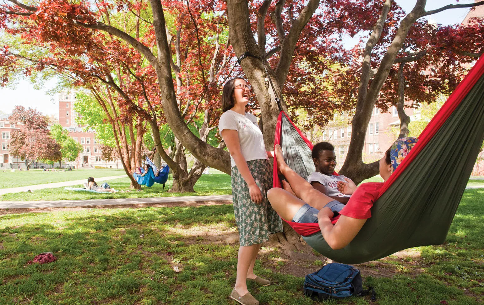 Three students relax in hammocks strung between trees with vibrant red leaves on a sunny day, while another student stands nearby laughing. In the background, other students lounge on the grass, and brick campus buildings are visible beyond the tree line.