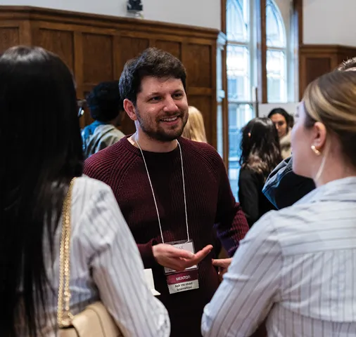 A male mentor wearing a name badge speaks with two students at an indoor networking event. The group stands in front of tall windows and wood-paneled walls, engaged in conversation in a warmly lit space.