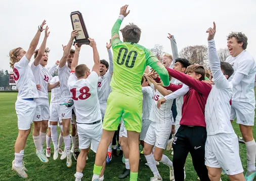 A college soccer team in white uniforms celebrates on a field as a player in a neon green goalkeeper outfit holds up a trophy. The team surrounds him with joyful expressions, raised arms, and cheering.