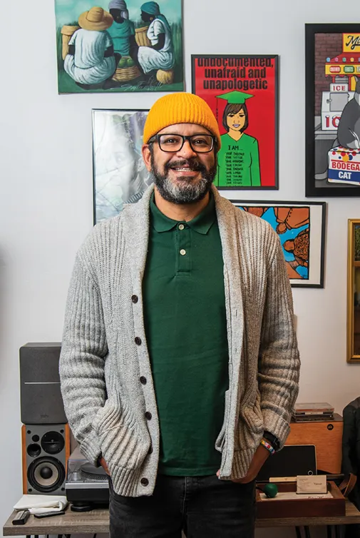 A man wearing a yellow knit hat, green polo shirt, and gray cardigan stands smiling in a colorful, art-filled room. Framed posters and illustrations cover the wall behind him, and audio equipment is visible in the lower part of the image.