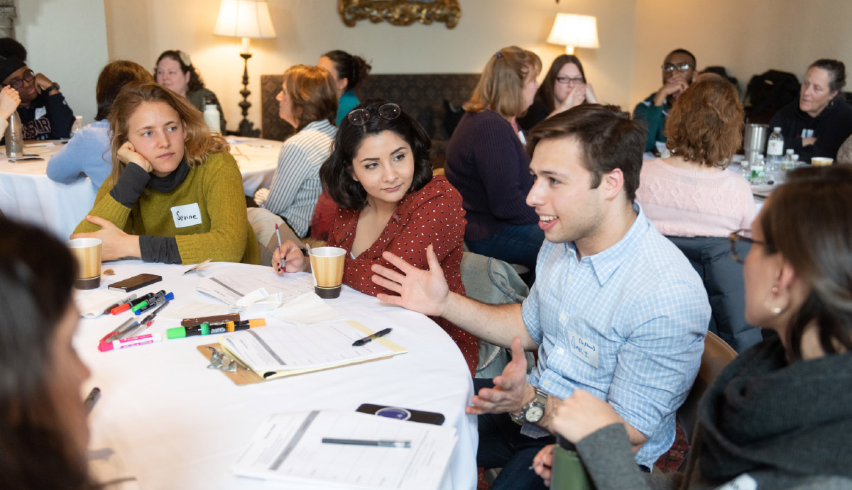 Students and faculty sit, in discussion, around tables with markers and paper in front of them.