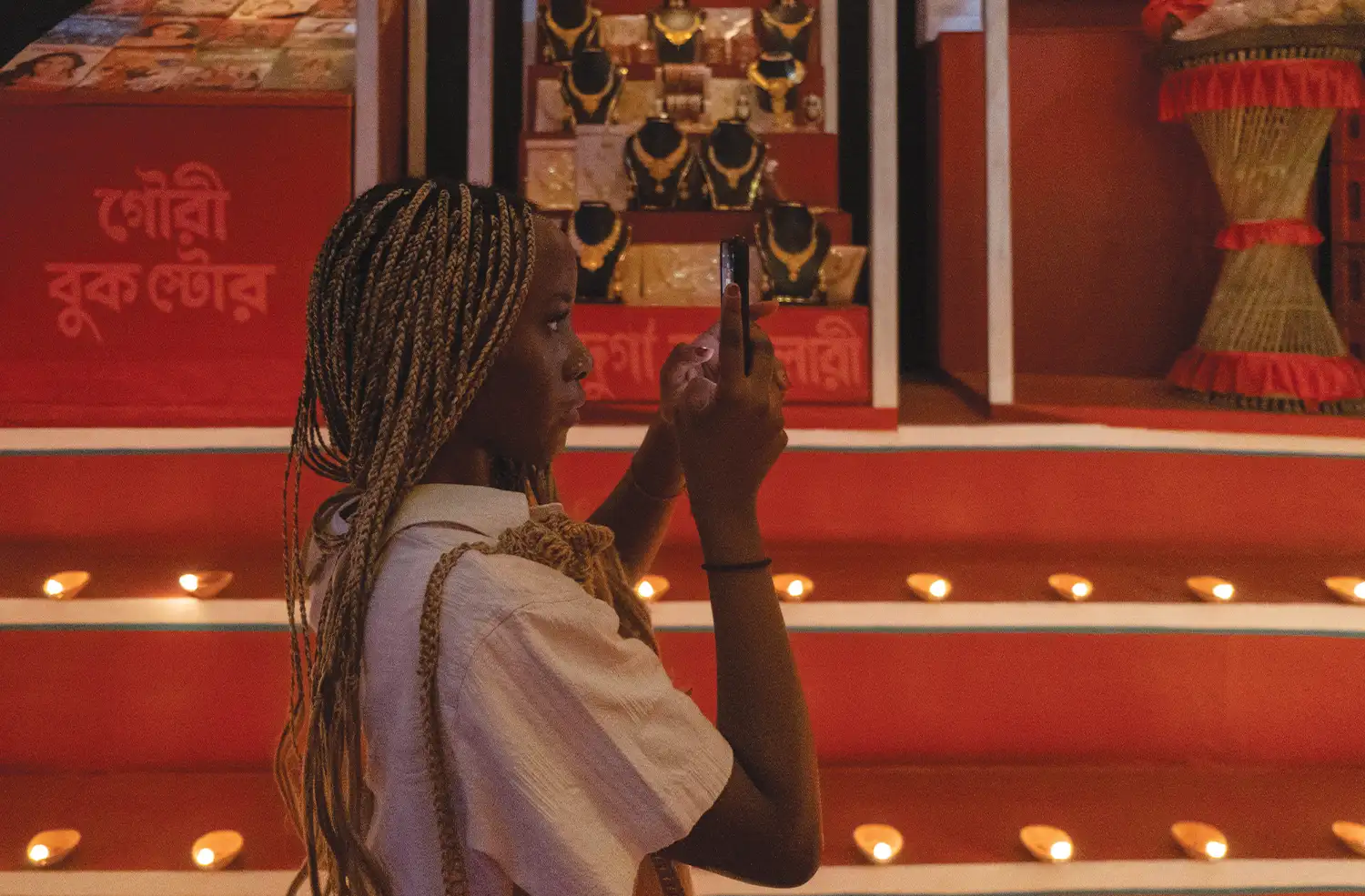 A student takes pictures on her phone of an exhibit with gold necklaces, red background, and candles.