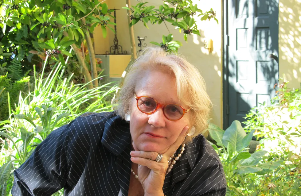 Portrait of Beth Dunlop wearing red-rimmed glasses, a dark pinstripe jacket, and pearl jewelry, seated outdoors in a lush green garden with a thoughtful pose, resting her chin on her hand.