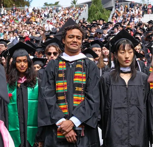 Group of students in black graduation gowns and caps standing together during commencement, with a crowd of attendees in the background.