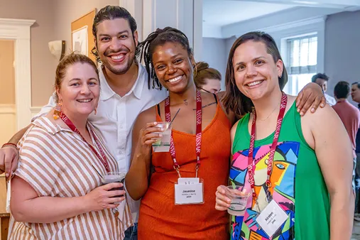 Four smiling alumni wearing nametags and holding drinks, gathered closely together at a reunion-style event indoors.