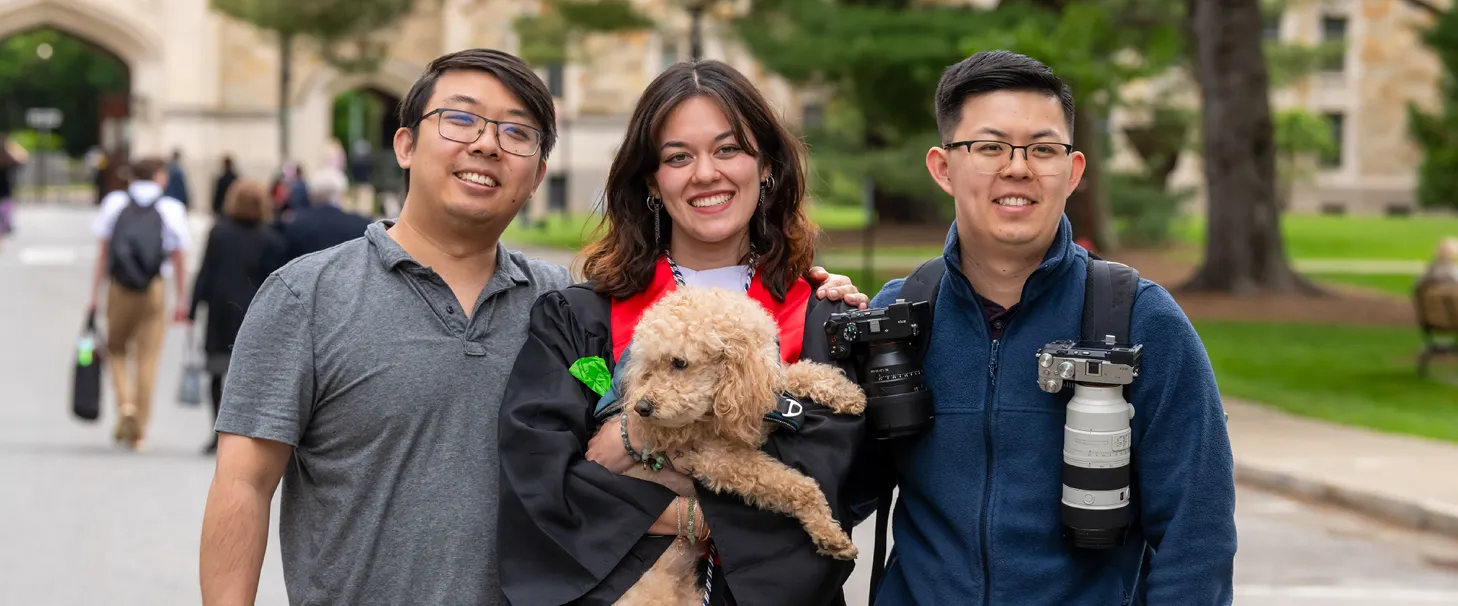 A graduate poses with two men while holding a dog.