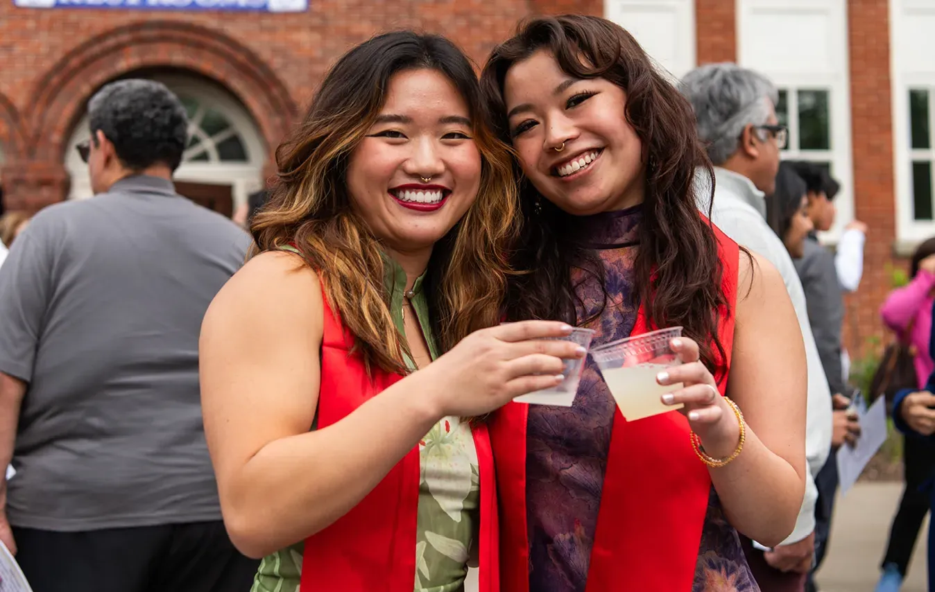 Two students, wearing red stoles to commemorate Asian and Pacific Islander identity, smile at the camera with drinks in their hands.
