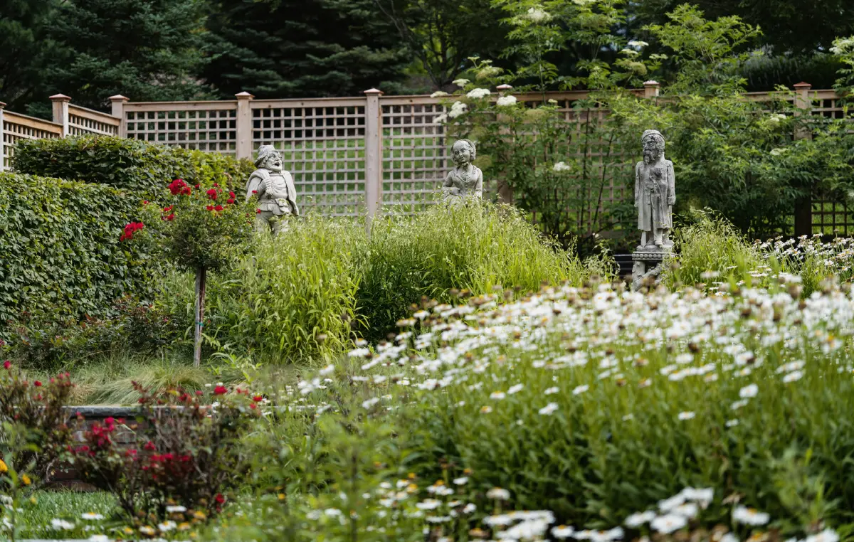 Landscape orientation outdoor photograph of a statue garden with three dark grey colored stone statues of what appear to be of historical figures or royalty figures; The statues are surrounded by overgrown green foliage, red roses, and a field of white daisy-like flowers; A light-colored faded dark brown wooden lattice fence is in the background situated amongst many trees in its nearby surroundings