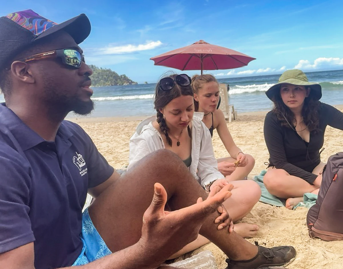 Four Vassar students sit on a beach in Trinidad and Tobago with a local tour guide.