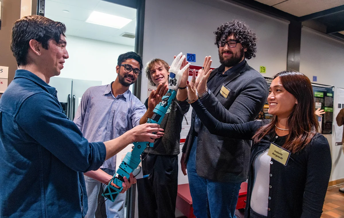 Students standing together, smiling, holding a robotic human arm.
