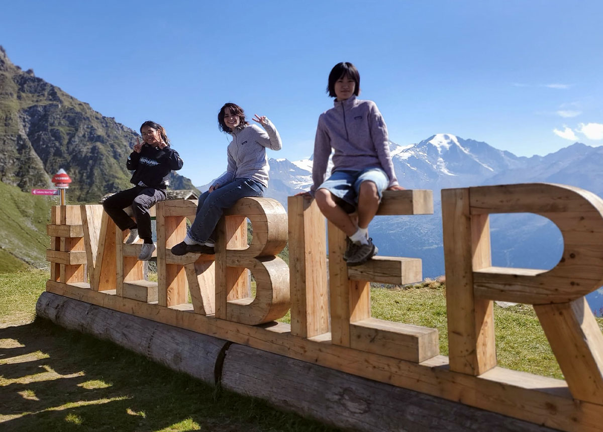Three students sitting on a wooden sculpture reading “#VERBIER”, smiling and posing for the picture.