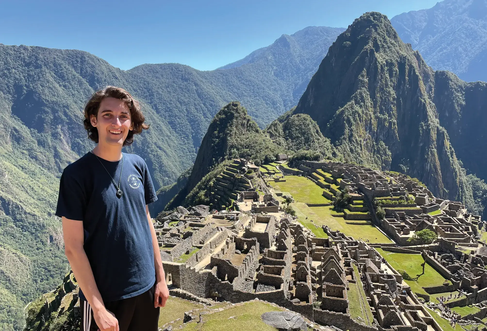 A smiling person stands on a ledge overlooking the stone terraces and ruins of Machu Picchu, with green mountains rising behind under a clear blue sky.