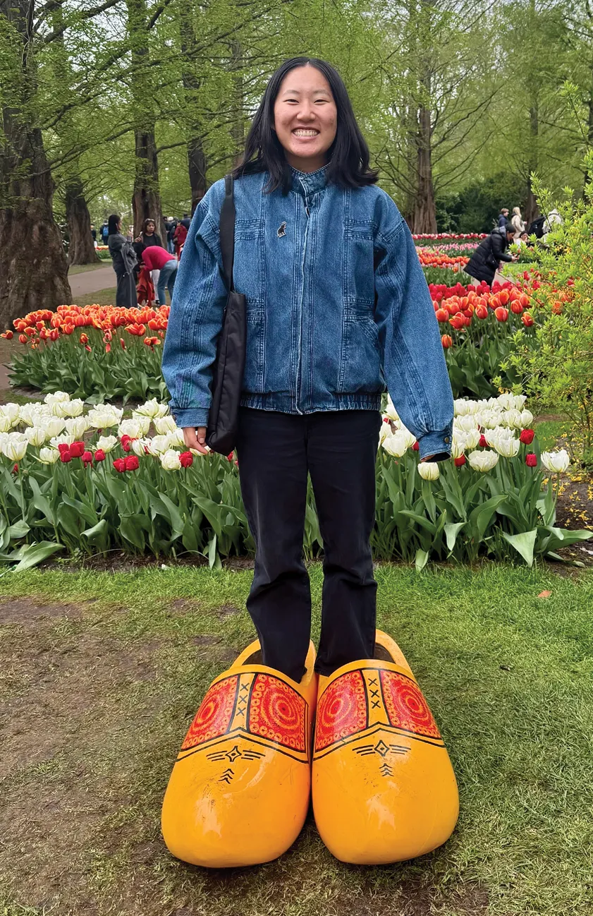 A person stands in a garden of colorful tulips wearing oversized decorative yellow wooden clogs, smiling with trees and other visitors in the background.