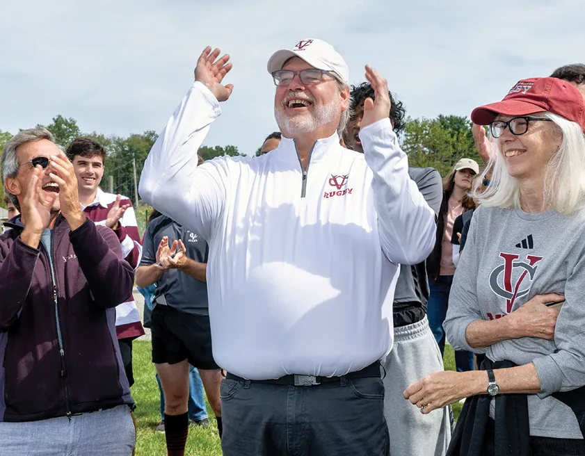 A group of people outdoors applaud and smile while a person wearing a white rugby shirt raises their hands enthusiastically, surrounded by teammates and supporters.