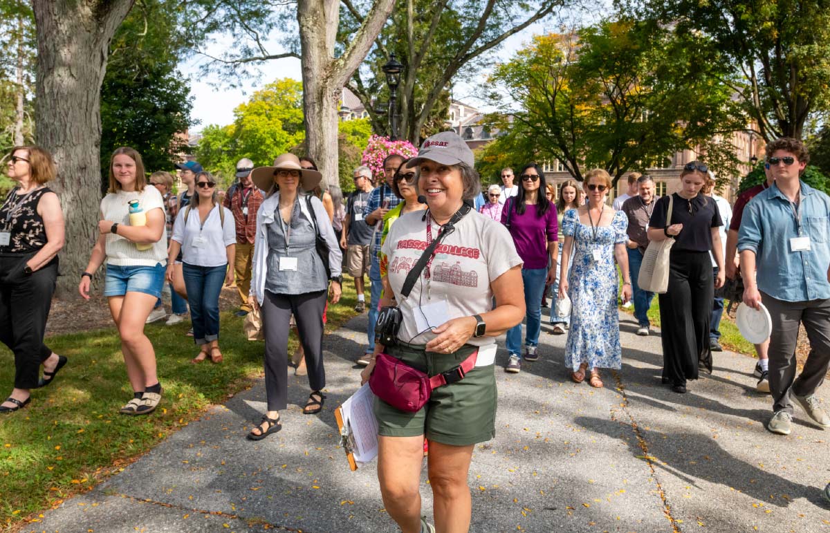 A woman in a Vassar t-shirt leads a large walking tour of families and students along a paved campus path on a sunny day.