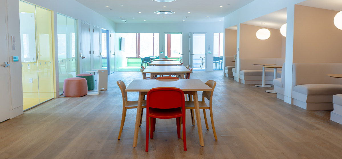 Spacious study area with light wood floors, various tables, chairs, and booths. Glass-enclosed rooms line the left, and spherical pendant lights hang over booths on the right.