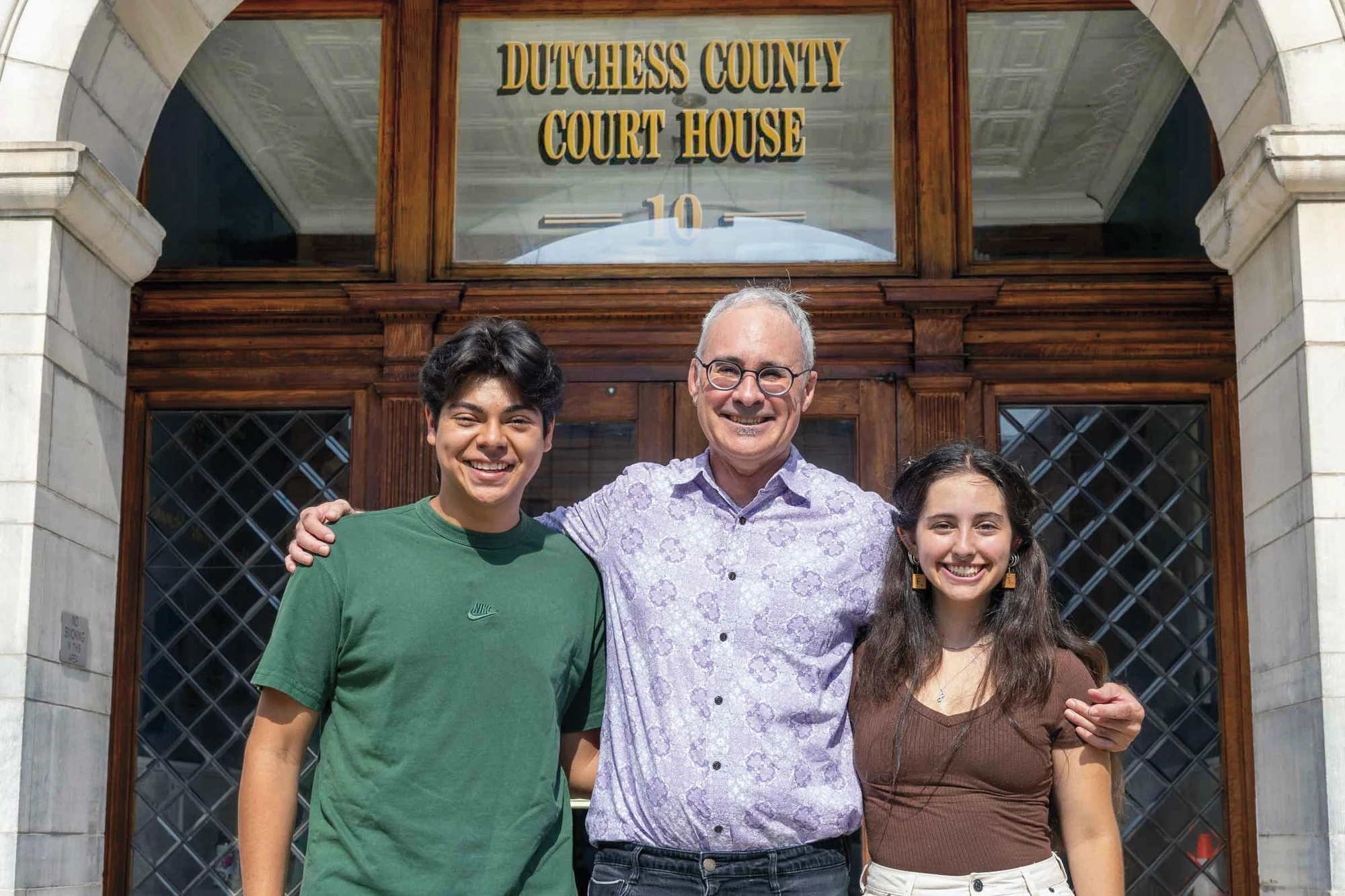 Three people stand with their arms around one another outside the Dutchess County Court House.