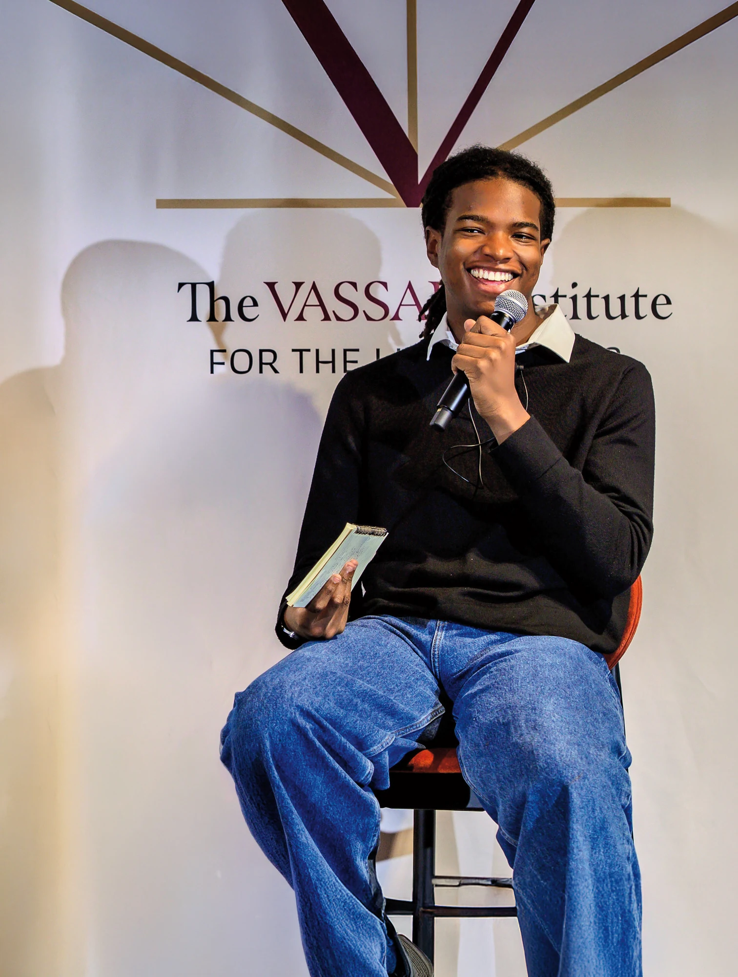A person with braided hair wearing a black sweater and blue jeans is sitting on a chair, smiling, holding a microphone in one hand and a notepad in the other, in front of a backdrop with the text "The Vassar Institute."