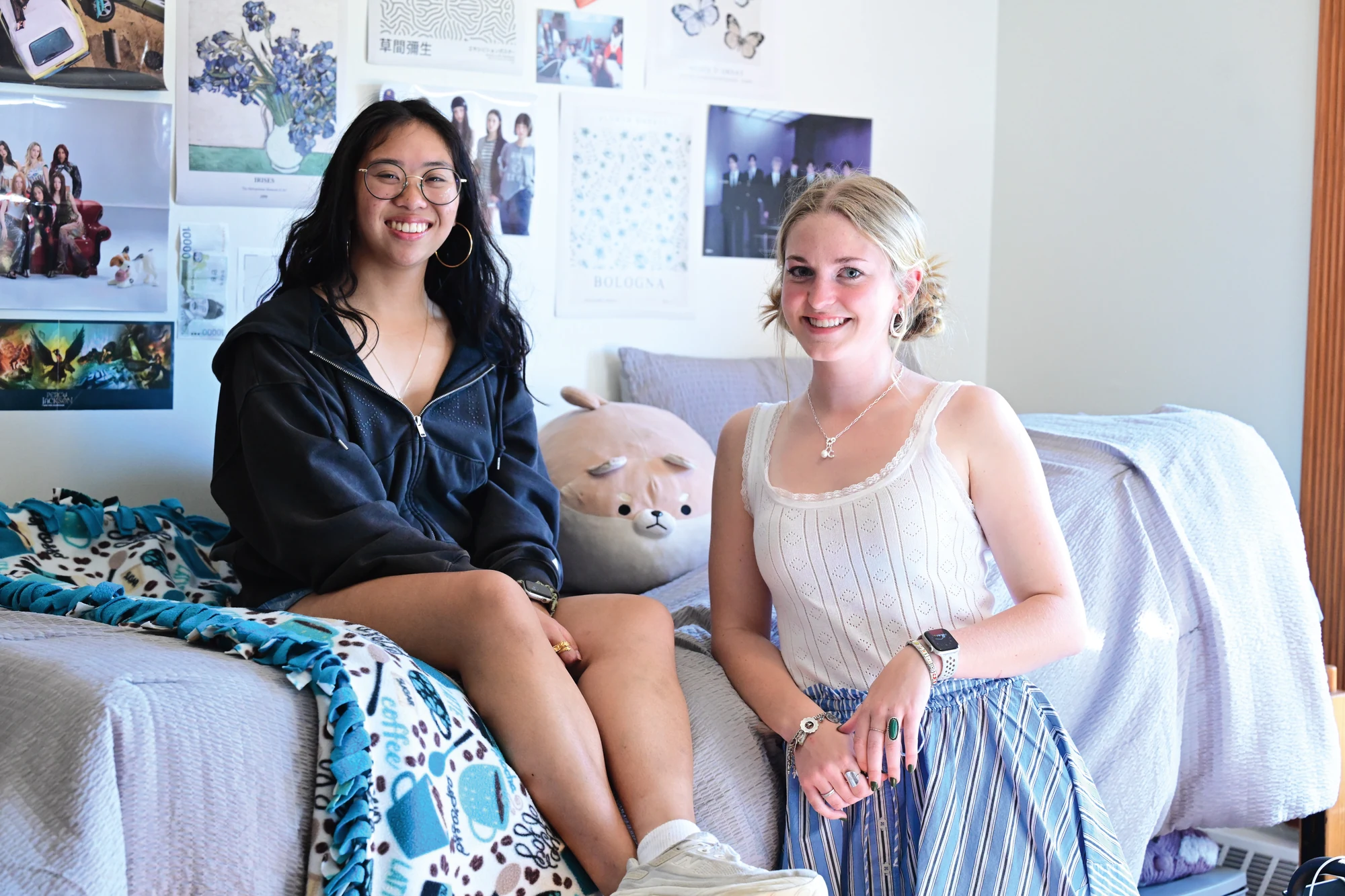 Two women sitting on a bed in a dorm smiling.