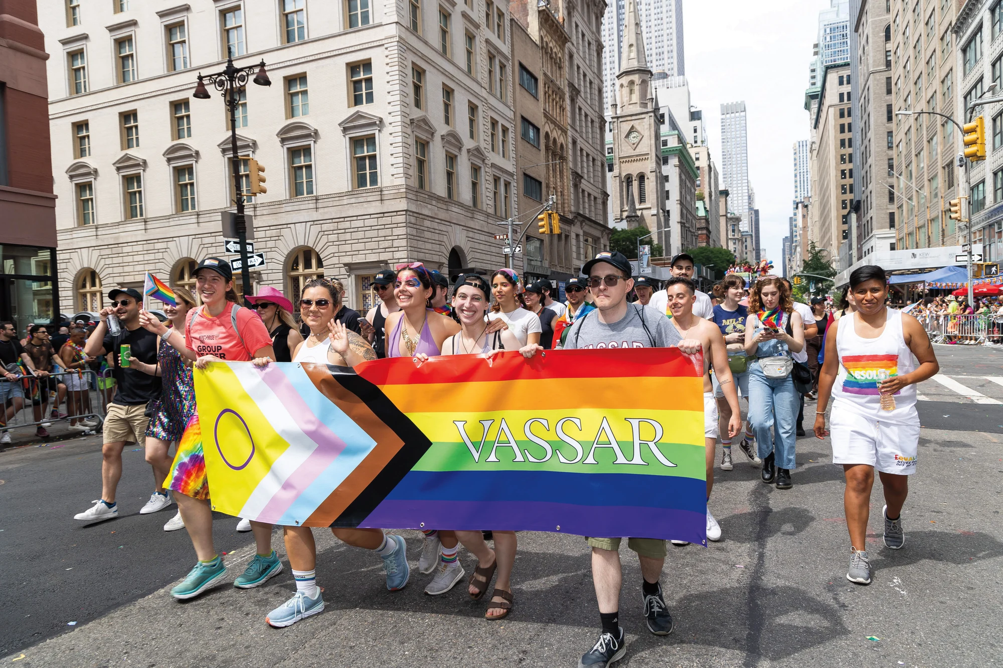 Vassar students march in a Pride parade carrying a rainbow banner and Pride flags.