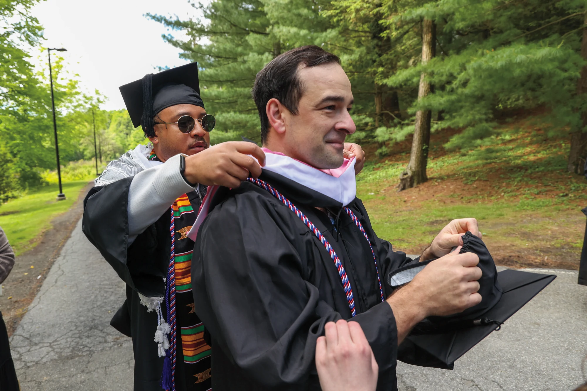 A Vassar graduate in cap and gown smiles while another graduate adjusts their academic hood.