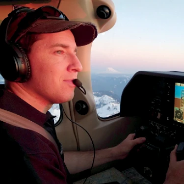 Max Fagin pilots an aircraft while wearing a headset, with snow-covered mountains visible outside.