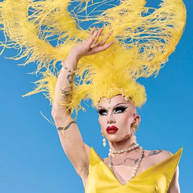 Sasha Velour poses in a yellow costume and dramatic feathered headpiece against a blue background.
