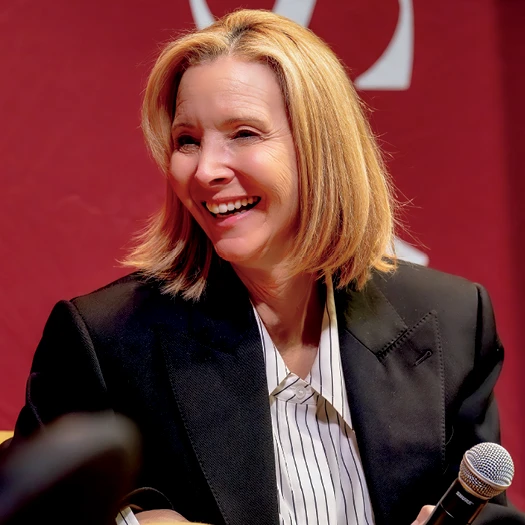 Lisa Kudrow laughs while holding a microphone during a public appearance.