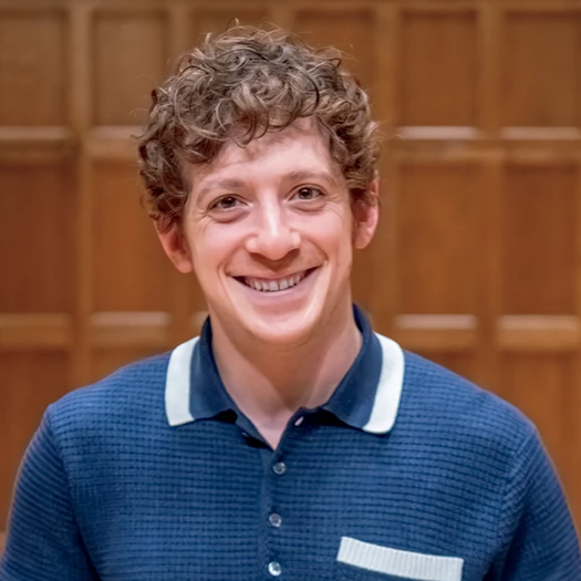 Ethan Slater smiles while standing in front of a wood-paneled wall.